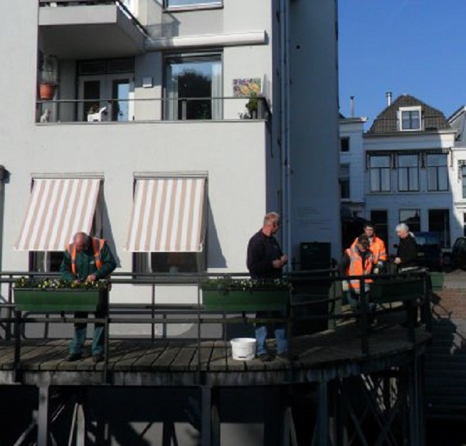 Gemeentewerklieden hangen bloembakken met winterviolen op. Ans kijkt toe